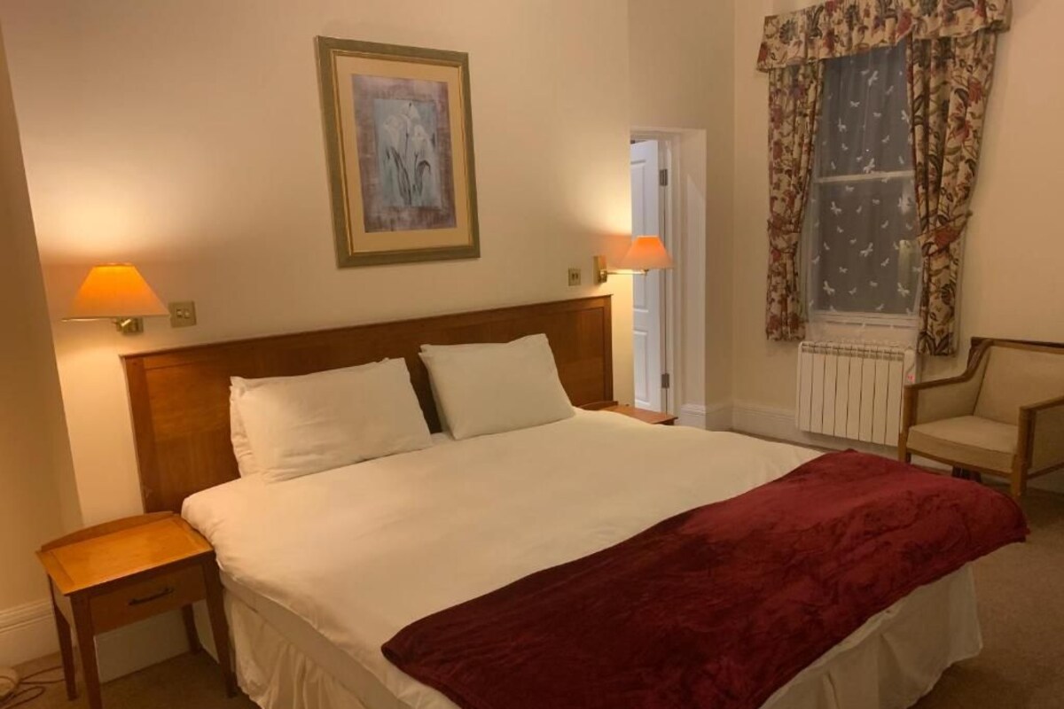 Une chambre d’hôtel avec un grand lit, deux lampes de chevet, une table de nuit, une chaise et une fenêtre avec des rideaux.