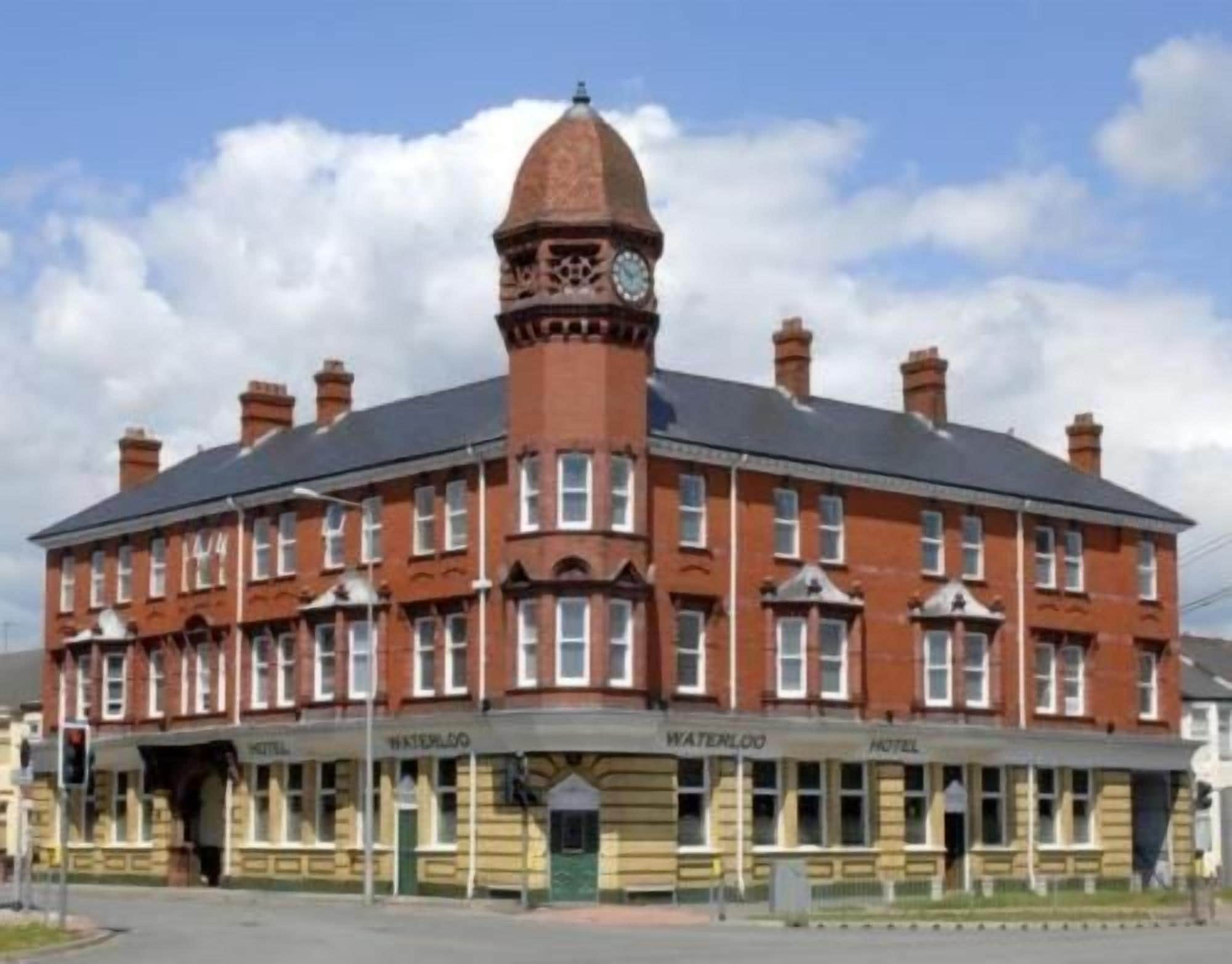 A red-brick building with a central tower and a clock. It has multiple windows and a sign that reads 'Waterloo Hotel.'