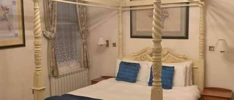 A four-poster canopy bed with a white frame and blue bedding, a bedside table, and a wall-mounted lamp.