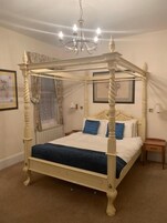 A four-poster canopy bed with a white frame and blue bedding, a bedside table, and a wall-mounted lamp.