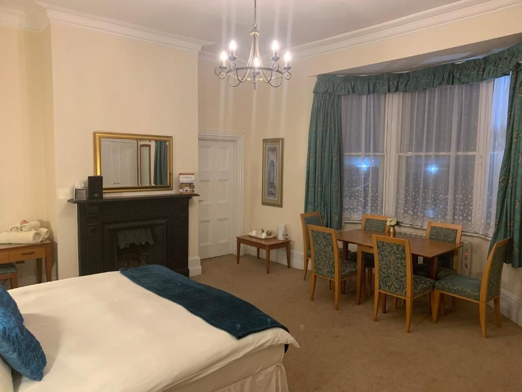 A hotel room with a bed, a fireplace, a dining table with chairs, and a large window with curtains.