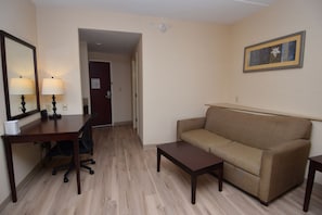 Una habitación de hotel con un escritorio de madera, un sofá color crema, una pequeña mesa de centro y un cuadro en la pared.