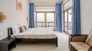 Une chambre d’hôtel avec un lit, une table de chevet, un canapé et un balcon avec des rideaux bleus.