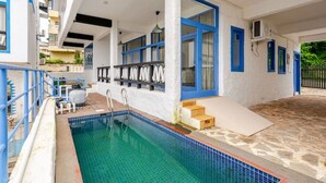 Une piscine avec un bord carrelé, un espace couvert doté d’un banc et un escalier menant à une porte.