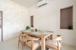 A dining area with a wooden table and chairs, an air conditioning unit, and two windows with blinds.