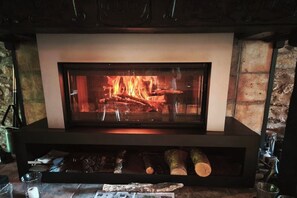 A fireplace with logs burning, a stone wall, and a metal shelf with items. - Renovated Cévennes-style house – heated pool in the forest (Les Plantiers)