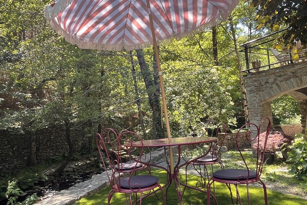 Eine Terrasse im Freien mit Tisch und Stühlen, umgeben von Grünpflanzen und einer Steinmauer.