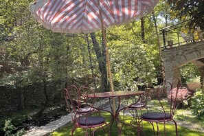 An outdoor patio with a table and chairs, surrounded by greenery and a stone wall. - Renovated Cévennes-style house – heated pool in the woods   (Les Plantiers)