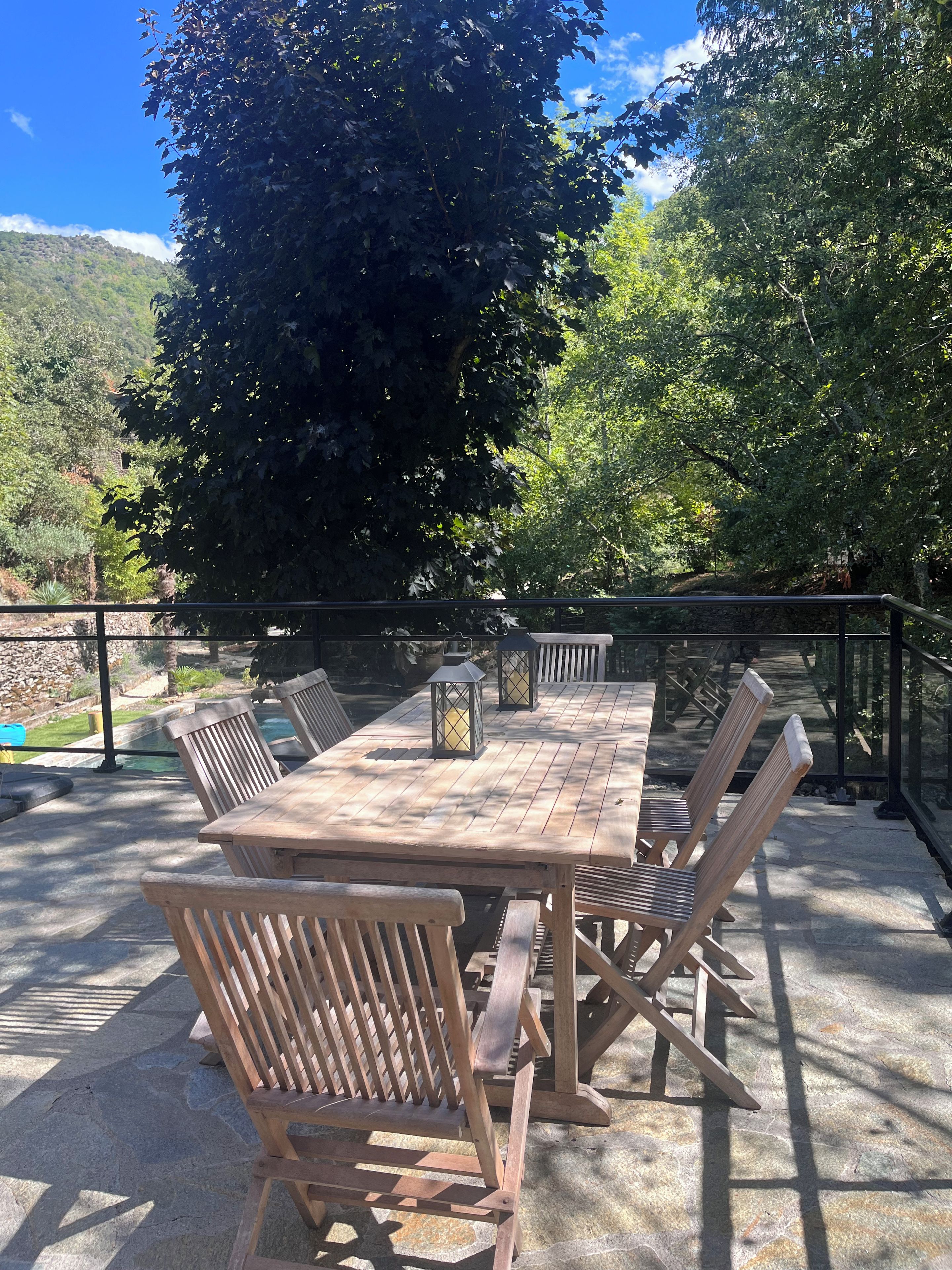 An outdoor patio with a wooden table and chairs, surrounded by stone flooring and a metal railing.