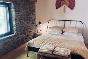 A bedroom with a stone wall, a bed with a metal frame, a bedside lamp, and a window. - Renovated Cévennes-style house – heated pool in the forest (Les Plantiers)