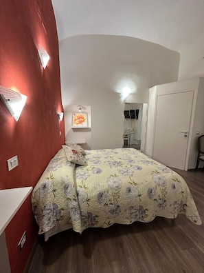 A bedroom with a floral bedspread, a red accent wall, and a white door. - Marcelina Vatican Rooms (Roma)