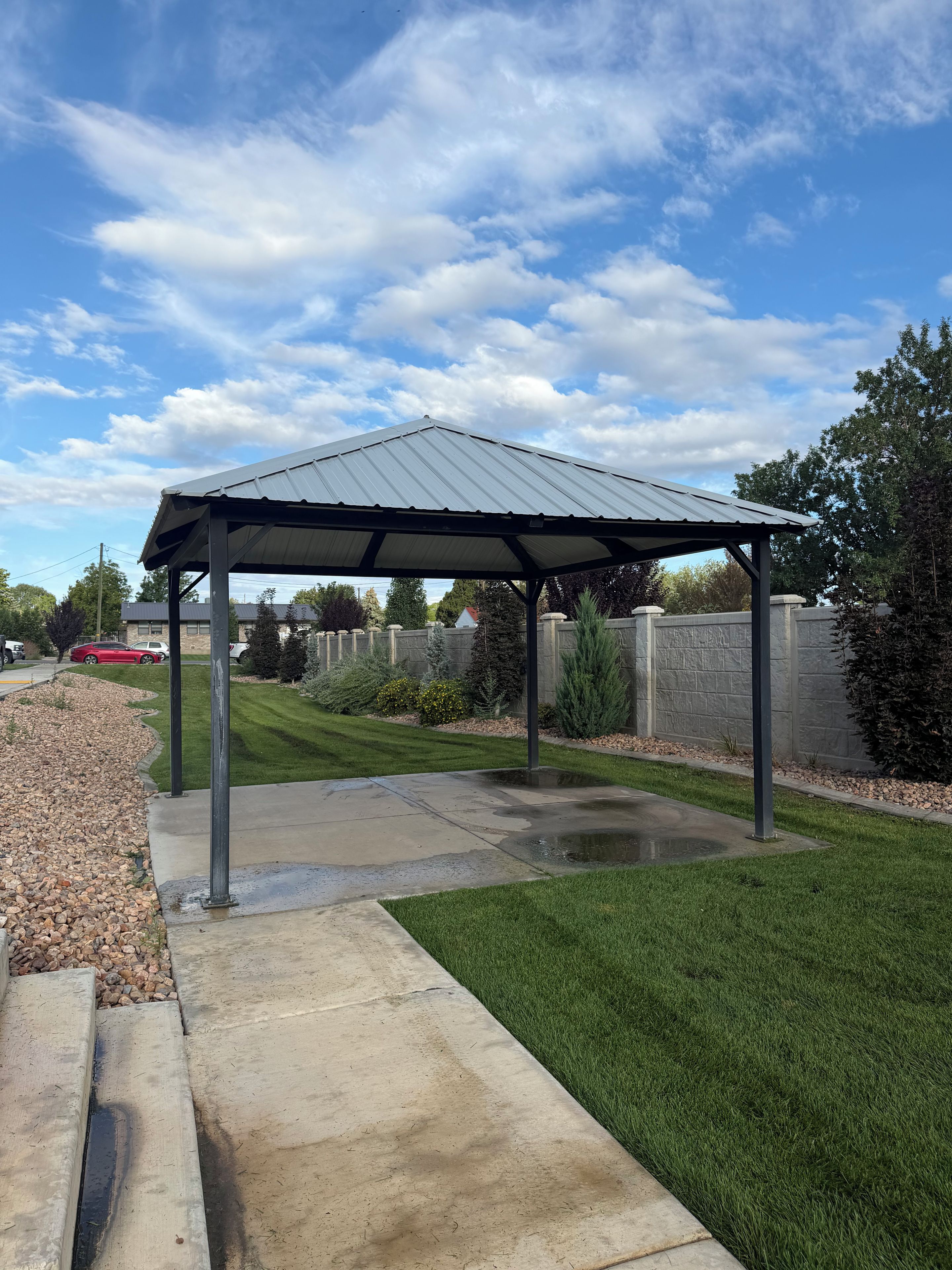 A covered patio area with a concrete floor and a metal roof, surrounded by a well-maintained lawn and stone walls.