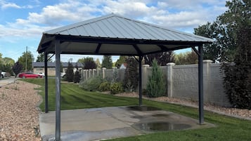 A covered patio area with a concrete floor and a metal roof, surrounded by a well-maintained lawn and stone walls.
