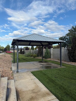Ein überdachter Patio-Bereich mit Betonboden und Metallüberdachung, umgeben von einem gepflegten Rasen und Steinmauern.