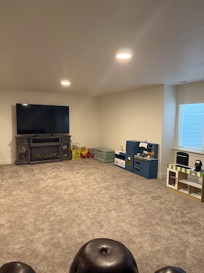A living room with a flat-screen TV, a fireplace, a play area with toys, and a small kitchenette.