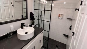Ein Badezimmer mit einem weißen Waschbecken, einem beleuchteten Spiegel, einer Duschkabine aus Glas und einer weißen Fliesenwand.