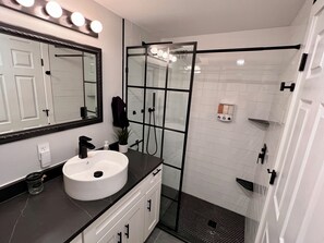 A bathroom with a white sink, a mirror with lights, a glass shower enclosure, and a white tiled wall. - Spacious and clean basement apartment minutes away from Roseau! (Roseau)