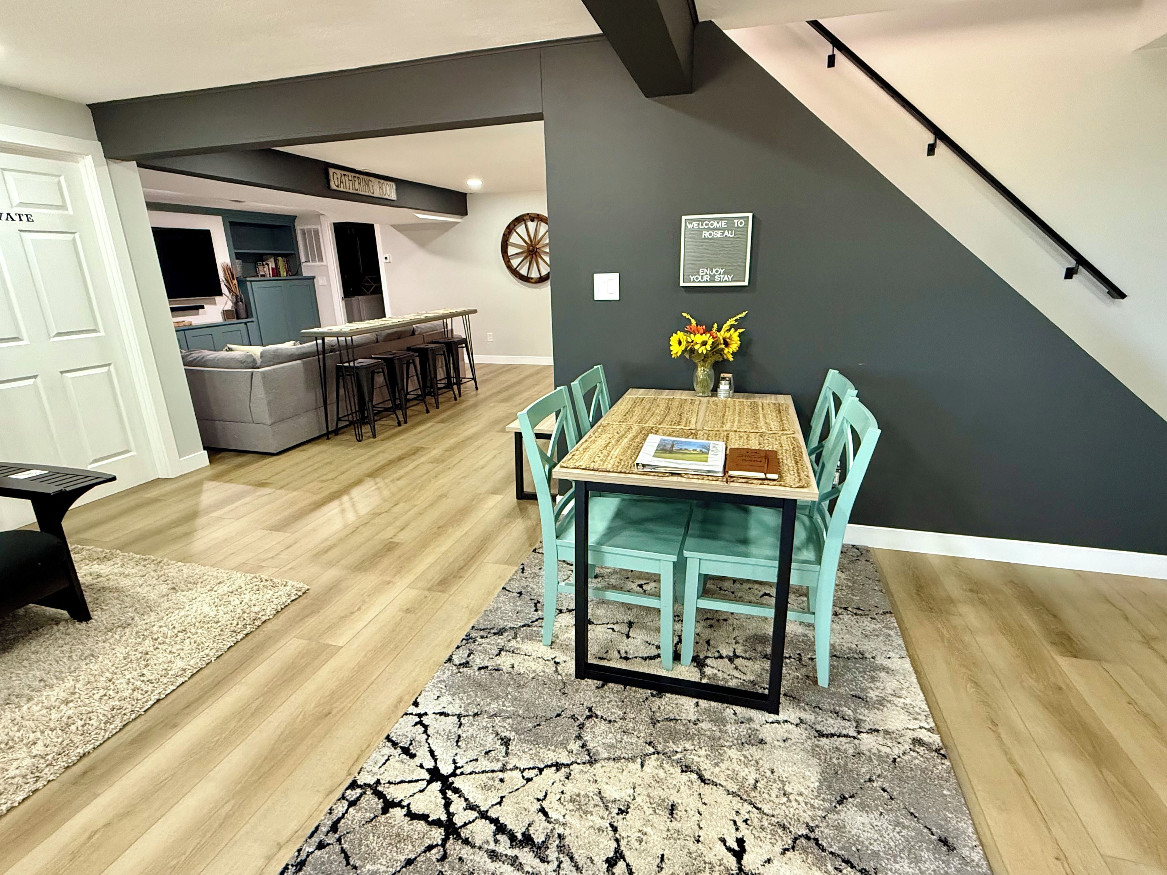 A dining area with a table and chairs, a rug, and a wall-mounted sign.