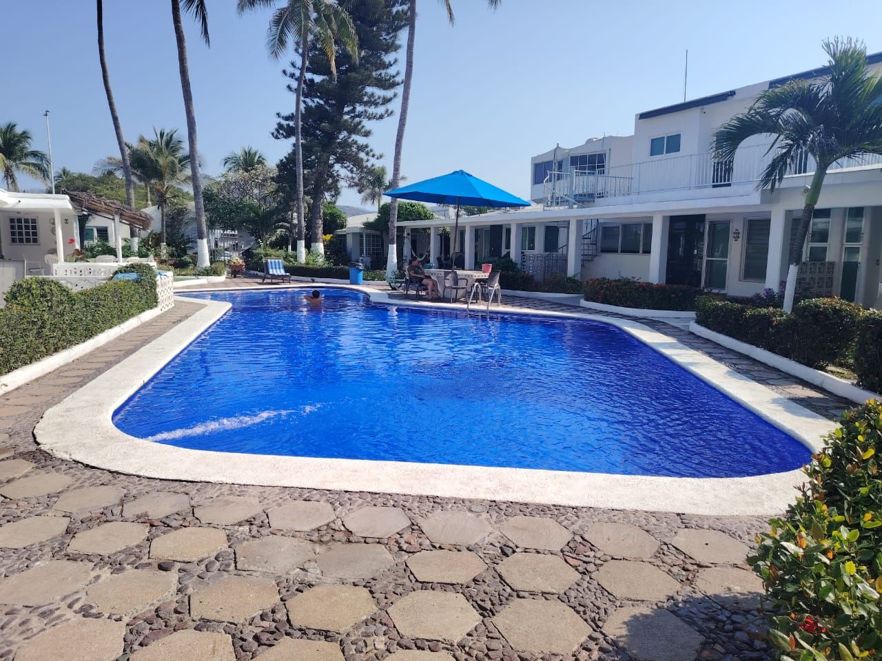 A swimming pool with a stone-tiled deck, surrounded by palm trees and a building with a balcony.