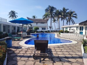 A swimming pool with a wooden bench in the foreground, surrounded by lounge chairs and a blue umbrella. - Casa Posada del Sol (Manzanillo)
