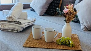 Un área de estar ordenada con una pila de toallas dobladas, dos tazas blancas, un jarrón con flores secas y uvas verdes, y un camino tejido sobre un sofá gris.