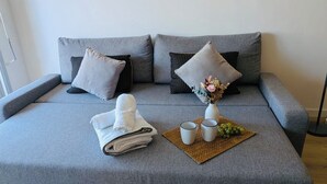 Un sofá gris con una toalla blanca doblada, dos tazas blancas, un jarrón pequeño con flores y un mantel tejido.