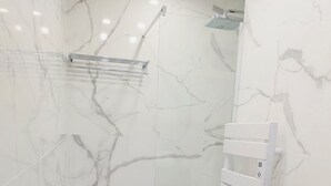 Un baño moderno con pared de ducha de mármol, cabina de ducha de vidrio, tocador de madera con lavamanos y dispensador de toallas.