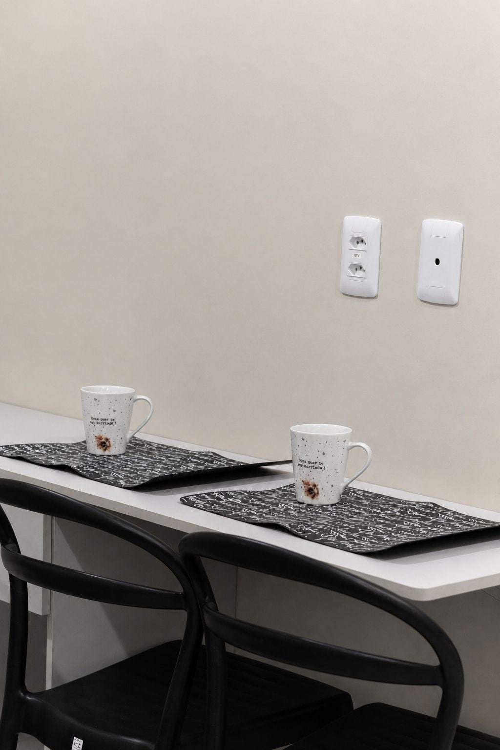 Una mesa de conferencias con dos tazas blancas sobre posavasos negros, una silla negra y dos enchufes en la pared.