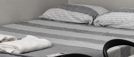 A single bed with striped bedding, a white towel, and a smartphone on the bed. There is a chair and a small table with a book or magazine.
