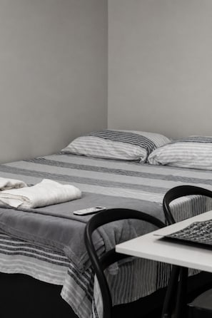 A single bed with striped bedding, a white towel, and a smartphone on the bed. There is a chair and a small table with a book or magazine.