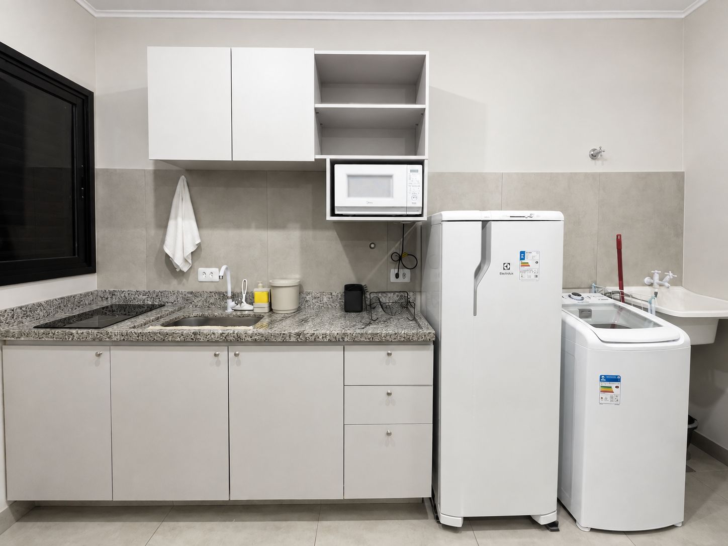 Una cocina compacta con armarios blancos, microondas, refrigerador, pileta y ventana.