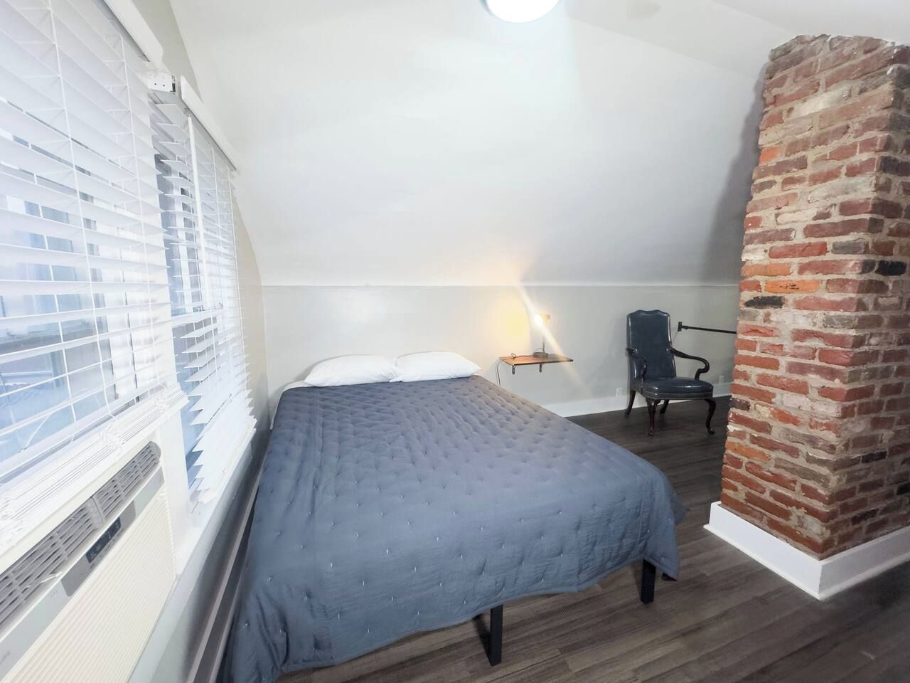 A bedroom with a bed, a chair, a brick wall, and a window with blinds.