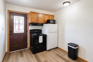A compact kitchen with a stove, microwave, refrigerator, and a trash bin.