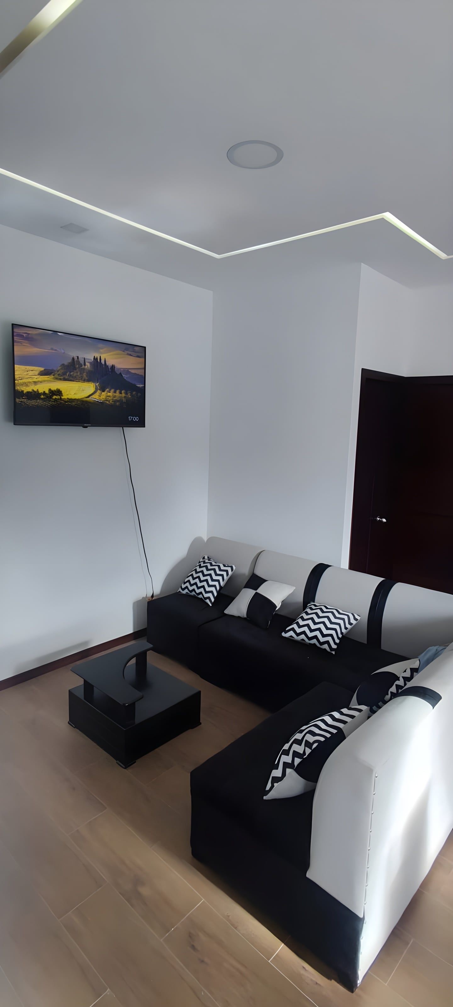 A modern living room with a flat-screen TV mounted on the wall, a black and white sofa, and a dark wooden door.