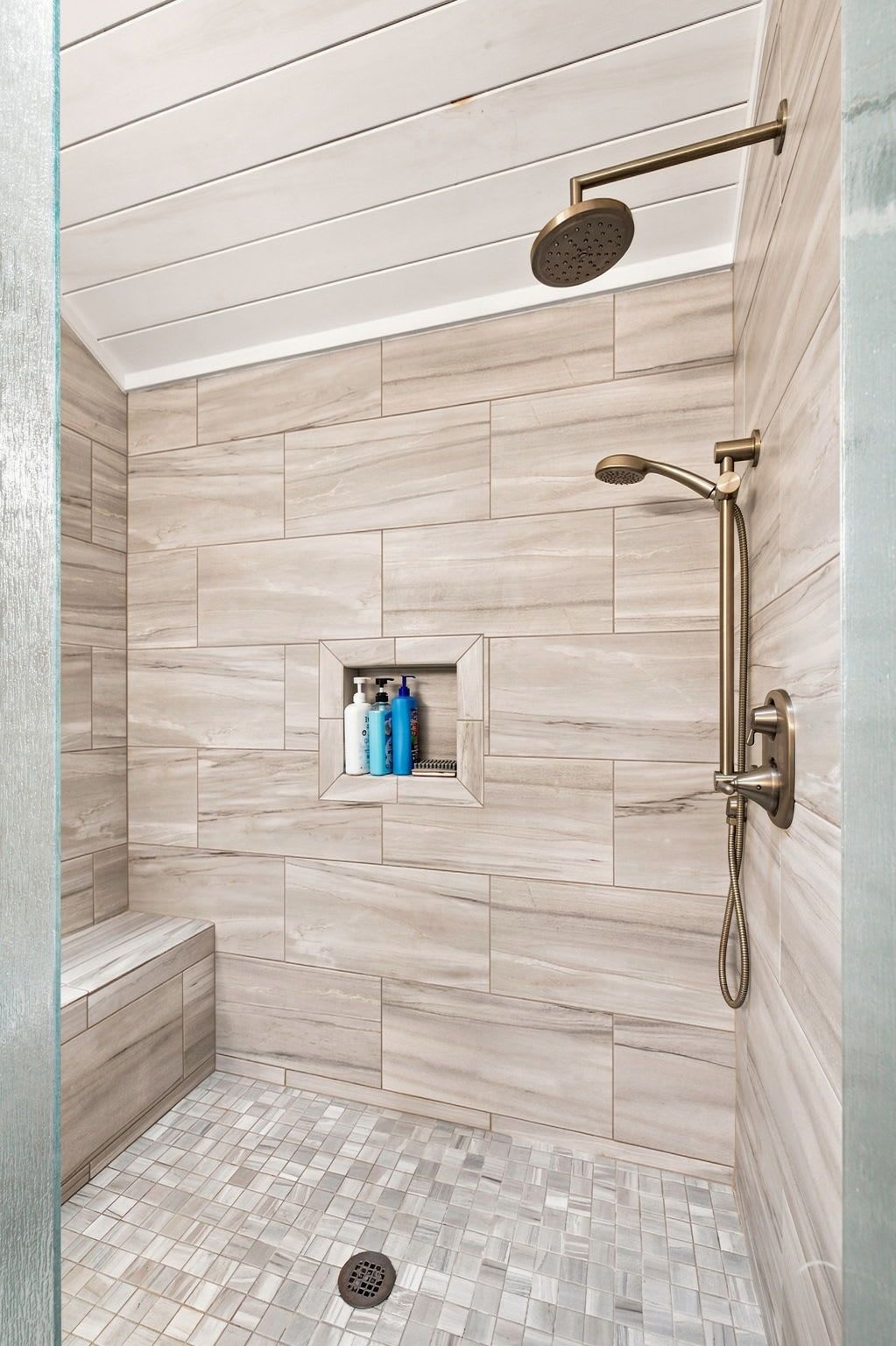 A shower with a handheld showerhead and a wall-mounted rain showerhead, a bench, and a shelf with toiletries.