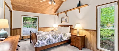 Ein Schlafzimmer mit Holzbalkendecke, einem Bett mit gemusterten Betttextilien, Nachttischen mit Lampen und großen Fenstern, die Blick auf Grünflächen bieten.