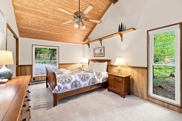 A bedroom with a wooden ceiling, a bed with patterned bedding, bedside tables with lamps, and large windows offering a view of greenery.