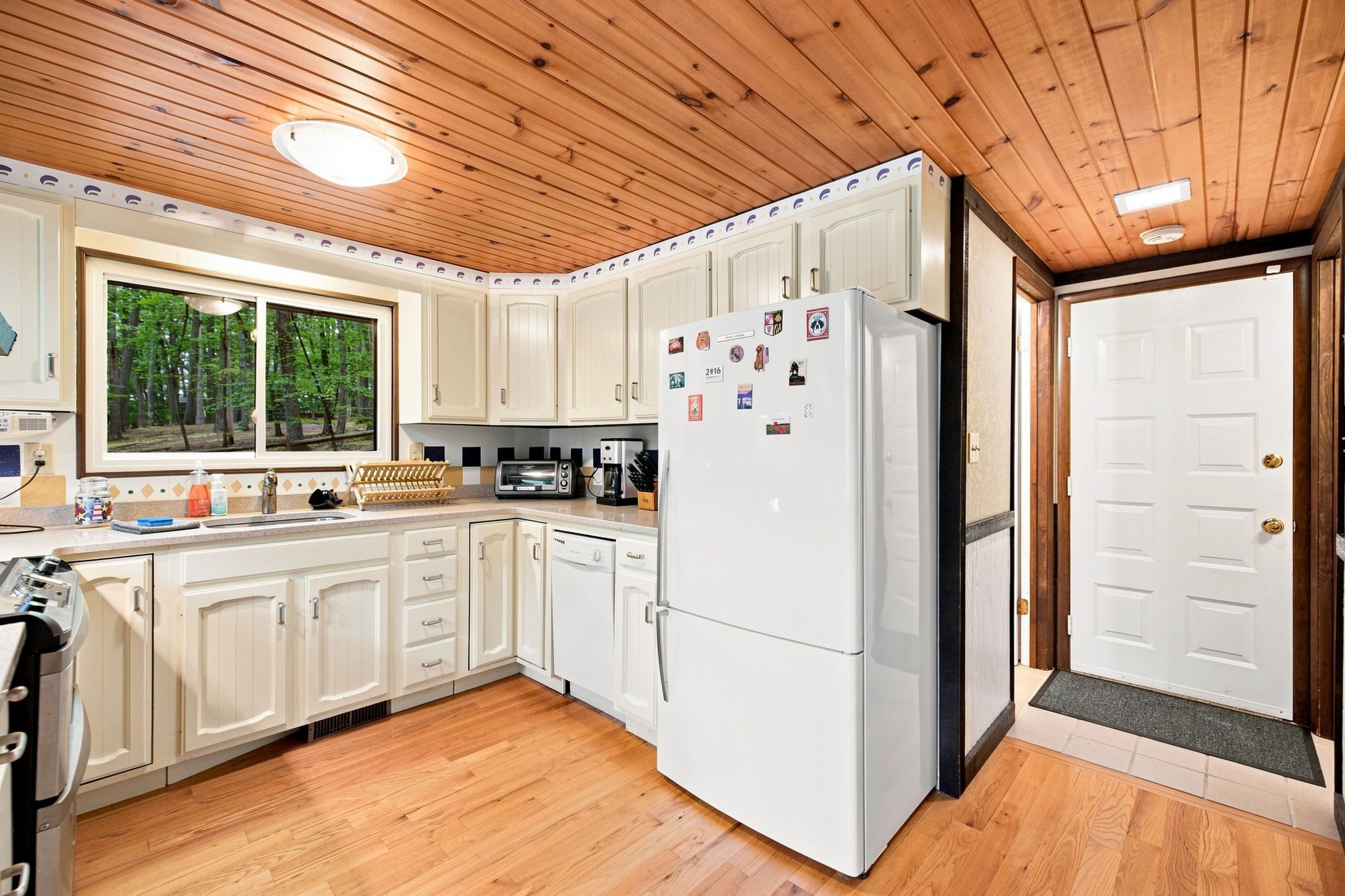 Una cocina con armarios de madera, un refrigerador y una ventana con vista a árboles.