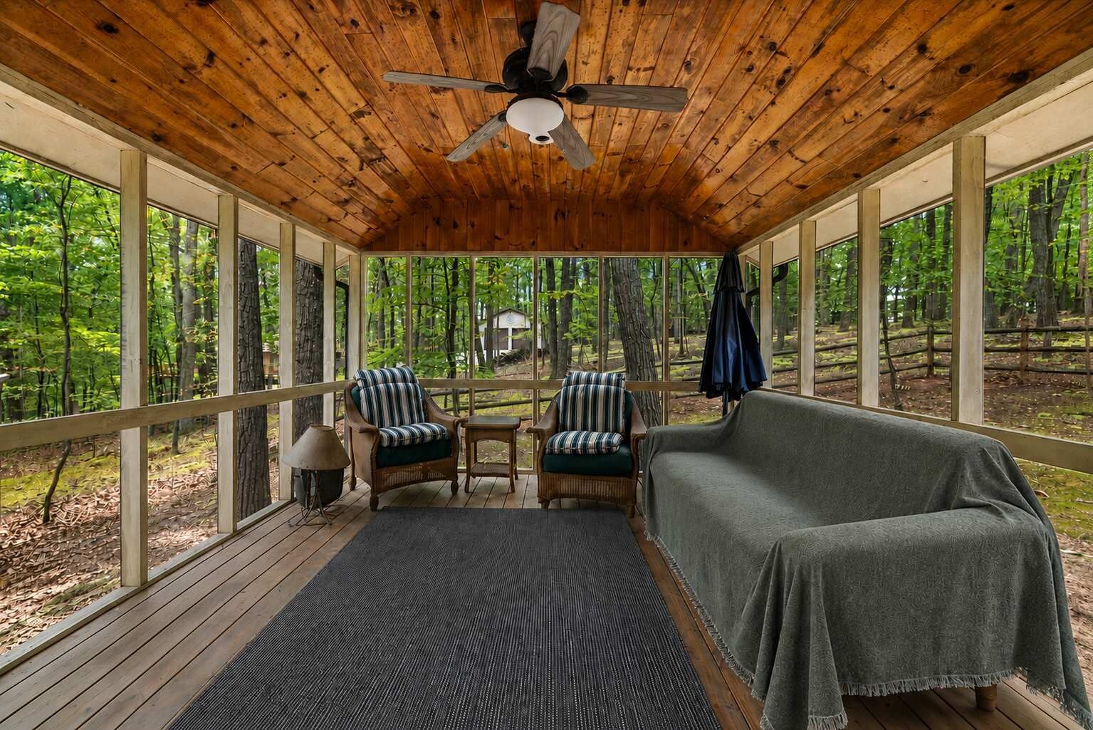 A wooden deck with a ceiling fan, two striped chairs, a sofa, and a view of a forest.