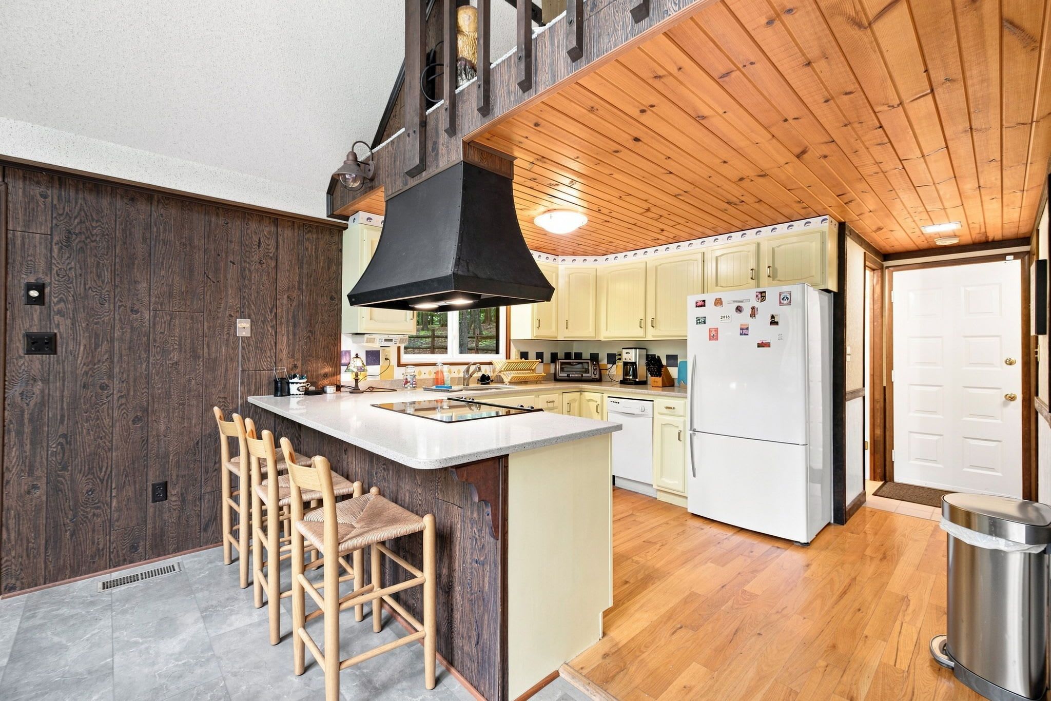 Een moderne keuken met een centraal eiland, houten barkrukken, een grote zwarte afzuigkap en een witte koelkast.