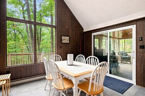 Un área de comedor con mesa y sillas, ventanas amplias con vista a árboles y una puerta corredera de vidrio que da a un espacio exterior.