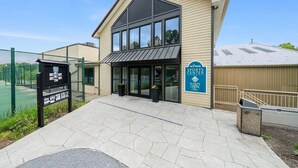 A sports center with a large glass entrance, a sign for 'The Woods Sports Center', and a fenced tennis court area. - Pet-Friendly with Treehouse, Deck in The Woods Resort (Hedgesville)
