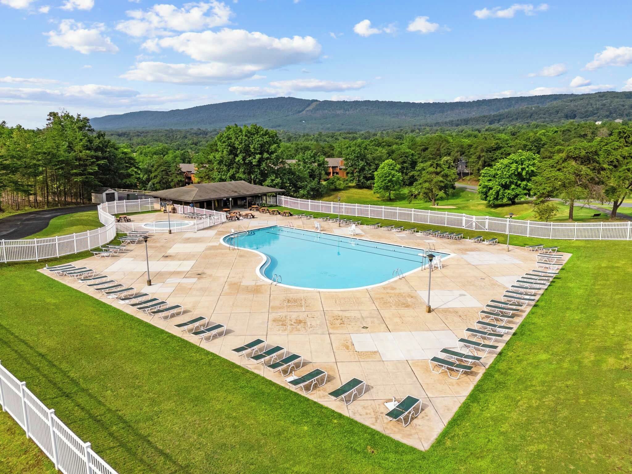 Una piscina con sillones de descanso y una zona cubierta, rodeada por una valla blanca, con vistas a colinas al fondo.
