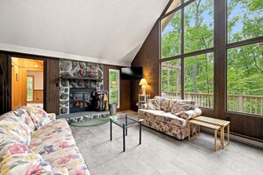 A cozy living room with a stone fireplace, a sofa, a coffee table, and a large window offering a view of the outdoors.