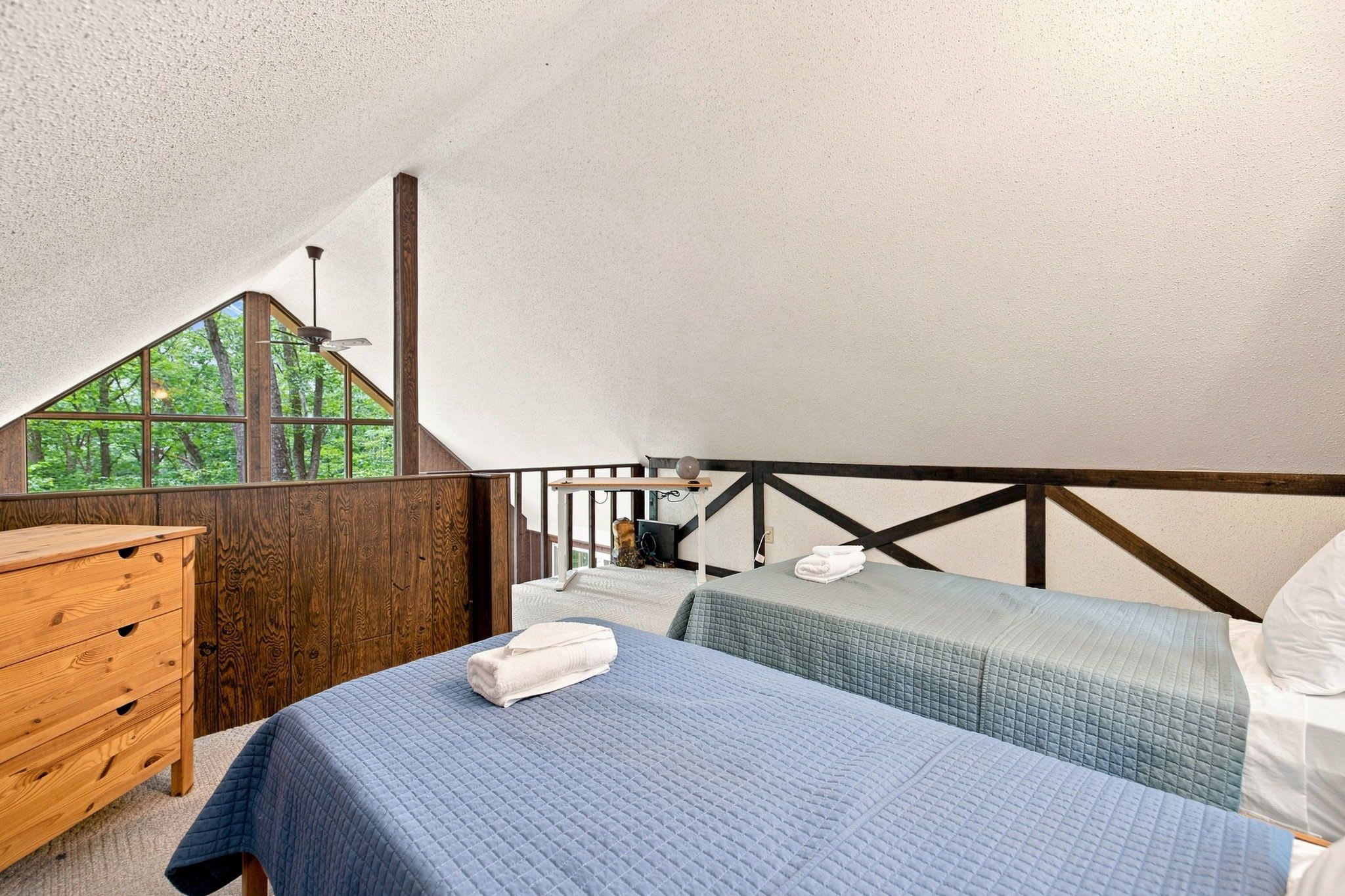 A bedroom with a sloped ceiling, wooden walls, a wooden dresser, two beds with blue bedding, and a window with a view of trees.
