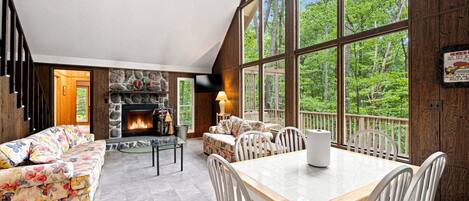 A spacious living room with a fireplace, a dining area with a table and chairs, and large windows offering a view of the outdoors.