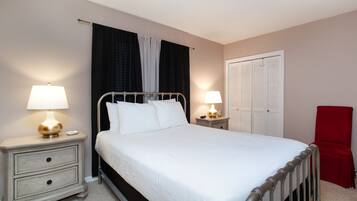 A neatly made bed with white linens, two bedside tables with lamps, a red chair, and a wardrobe with louvered doors.