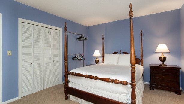 A four-poster bed with a wooden frame and white bedding, a bedside table with a lamp, and a clothing rack against the wall. - Smoky Laurel Cottage - Mountain Views & Fireplace! (Gatlinburg)