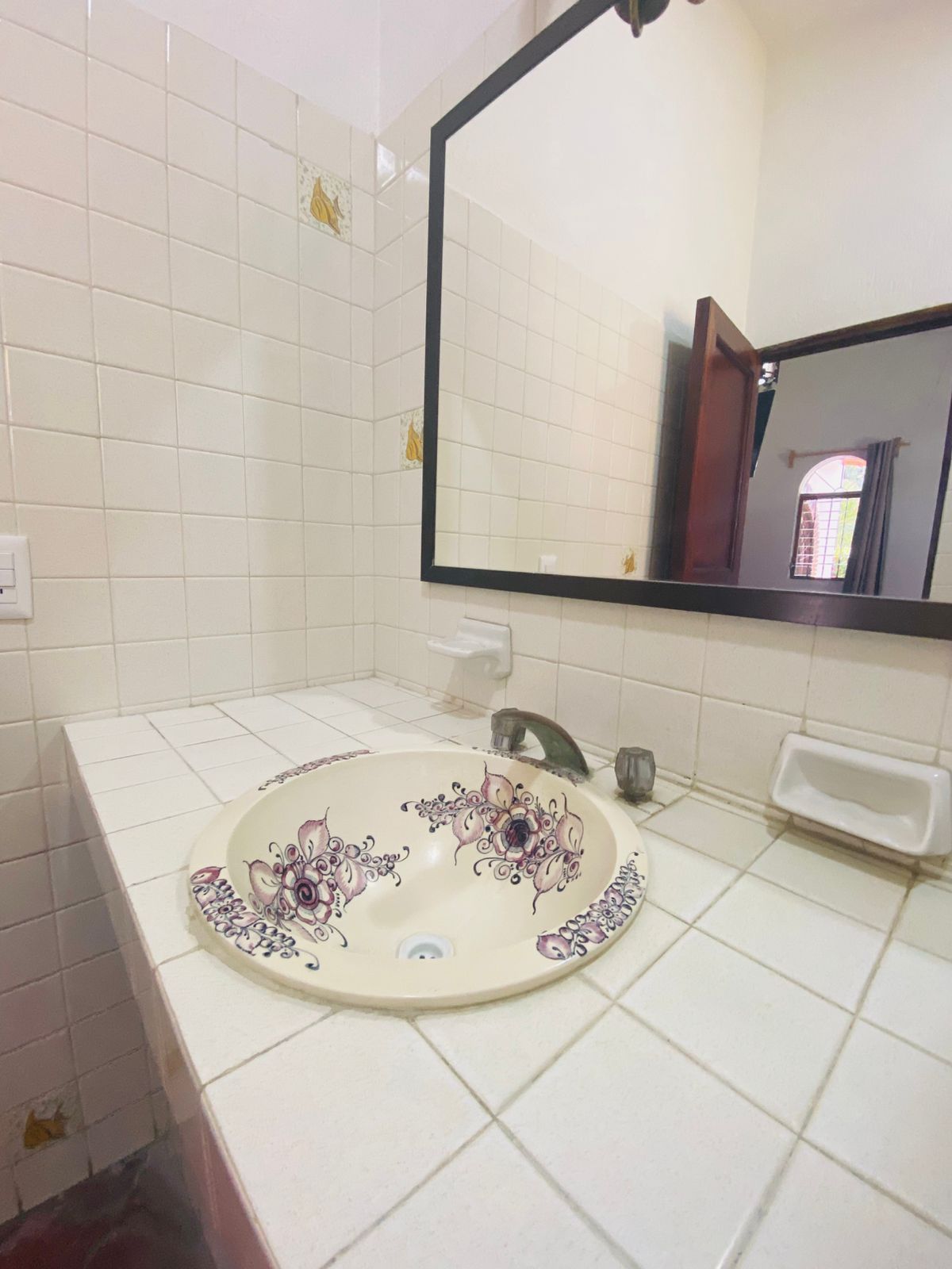 Un bany amb paret de rajoles blanques, un lavabo rodó amb motius florals, un mirall i una finestra amb vista.