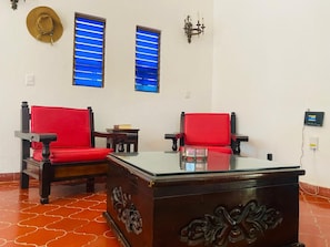 A room with two red upholstered chairs, a glass-top table, and a decorative cabinet. There are two windows with blue blinds, a wall-mounted light fixture, and a wall socket. - Silvia's House (Manzanillo)